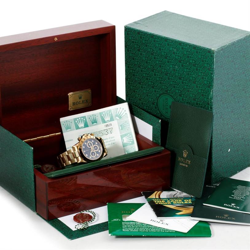The image shows a Rolex Daytona Men's Yellow Gold Black Dial Men's Yellow Gold Black Dial watch in a wooden presentation box, alongside certificates, booklets, and an outer green box.