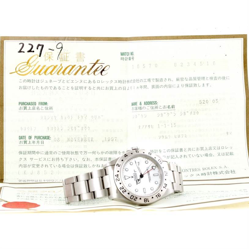 The image shows a Rolex Explorer 16570 Men
s Stainless Steel White Dial 16570 Men
s Stainless Steel White Dial watch laid flat on its side, with its warranty certificate displayed in the background.