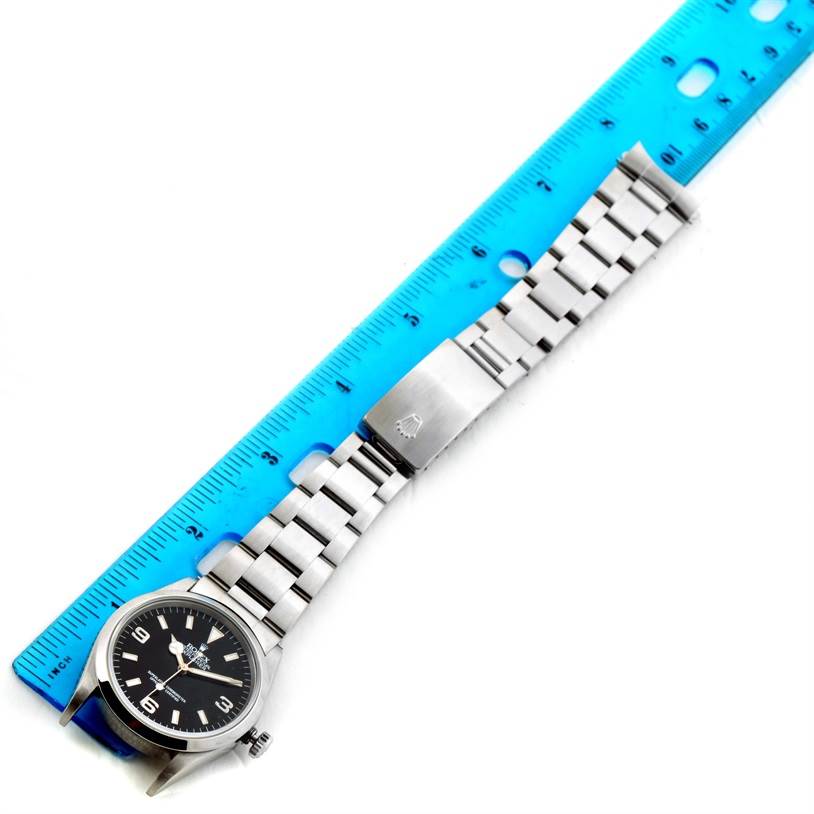 The Rolex Explorer 14270 Men's Stainless Steel Black Dial 14270 Men's Stainless Steel Black Dial watch is shown from an angled top view, displaying the face, bracelet, and clasp beside a blue ruler.