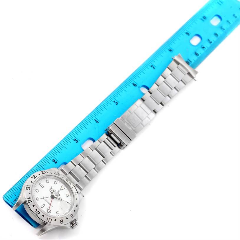 The Rolex Explorer 16570 Men
s Stainless Steel White Dial 16570 Men
s Stainless Steel White Dial watch is shown lying flat next to a blue ruler, displaying its face, bezel, and metal bracelet.