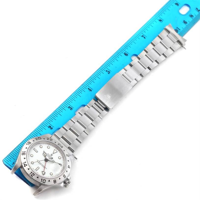 The Rolex Explorer 16570 Men's Stainless Steel White Dial 16570 Men's Stainless Steel White Dial watch is shown from a slanted top-down angle, displaying the face and the metal bracelet against a blue ruler.