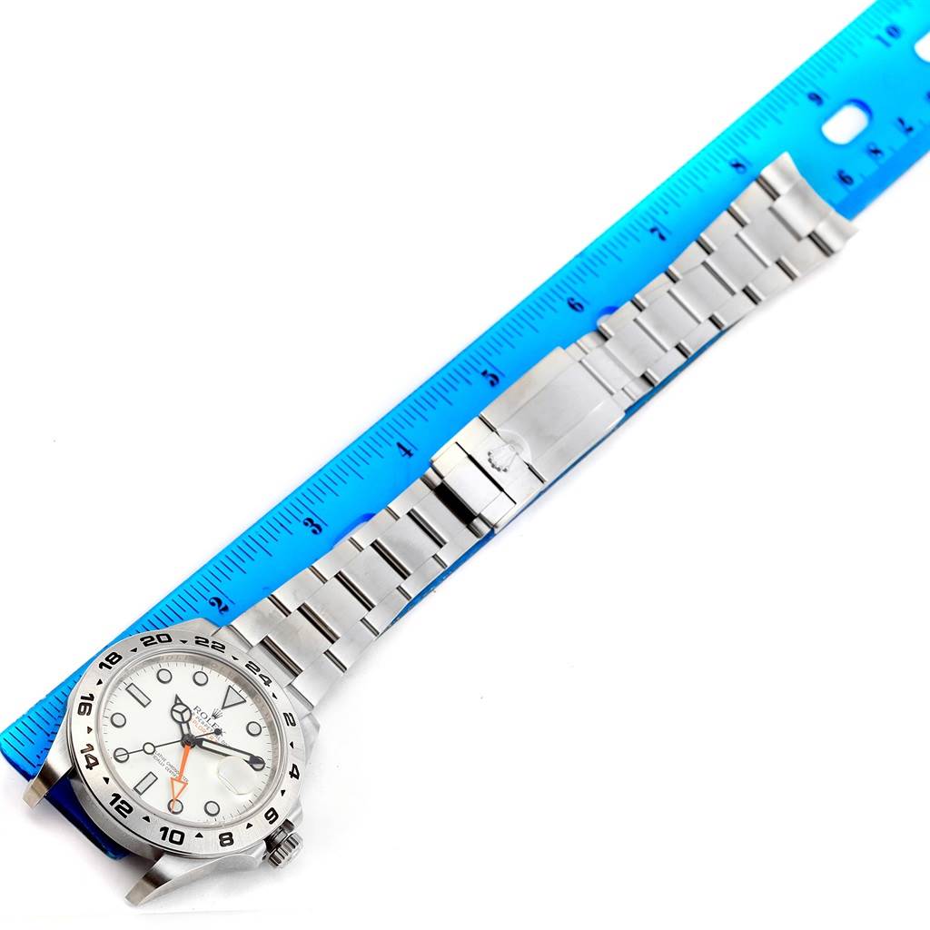 The image shows a top-down view of a Rolex Explorer 216570 Men's Stainless Steel White Dial 216570 Men's Stainless Steel White Dial watch with its stainless steel band next to a blue ruler.