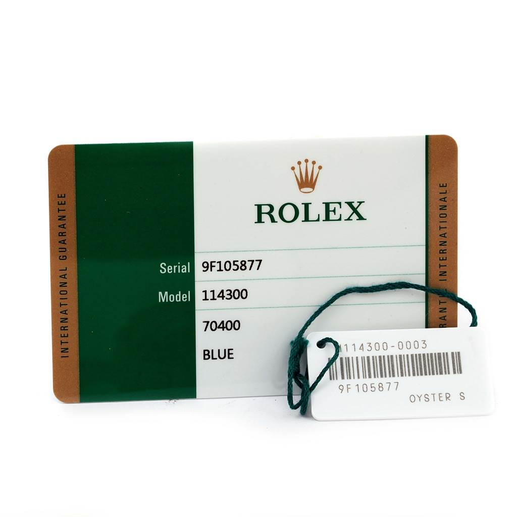 The image shows the warranty card and tag for a Rolex Explorer 114300 Men's Stainless Steel Blue Dial 114300 Men's Stainless Steel Blue Dial model, featuring the serial number, model number, and color.