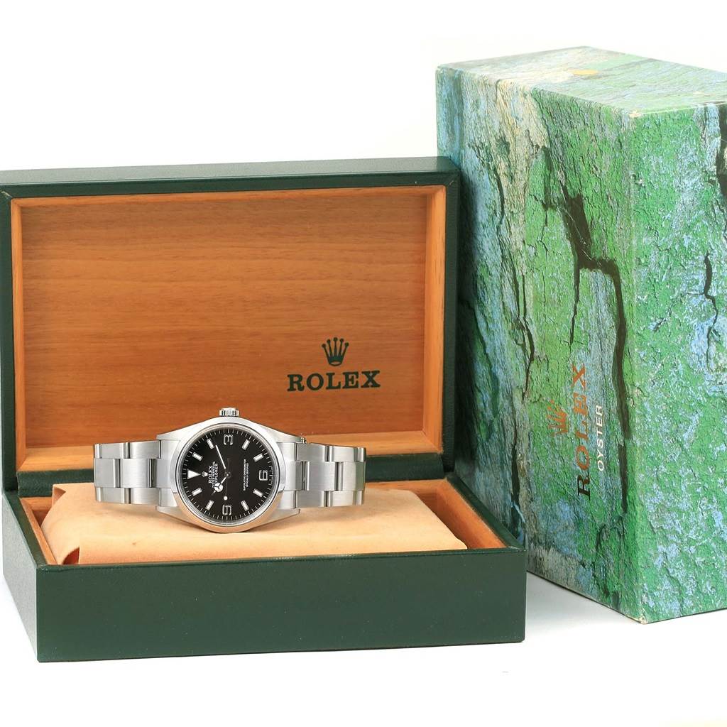 The image shows a Rolex Explorer 114270 Men's Stainless Steel Black Dial 114270 Men's Stainless Steel Black Dial watch with a black dial, presented in an open display box next to its green outer packaging.