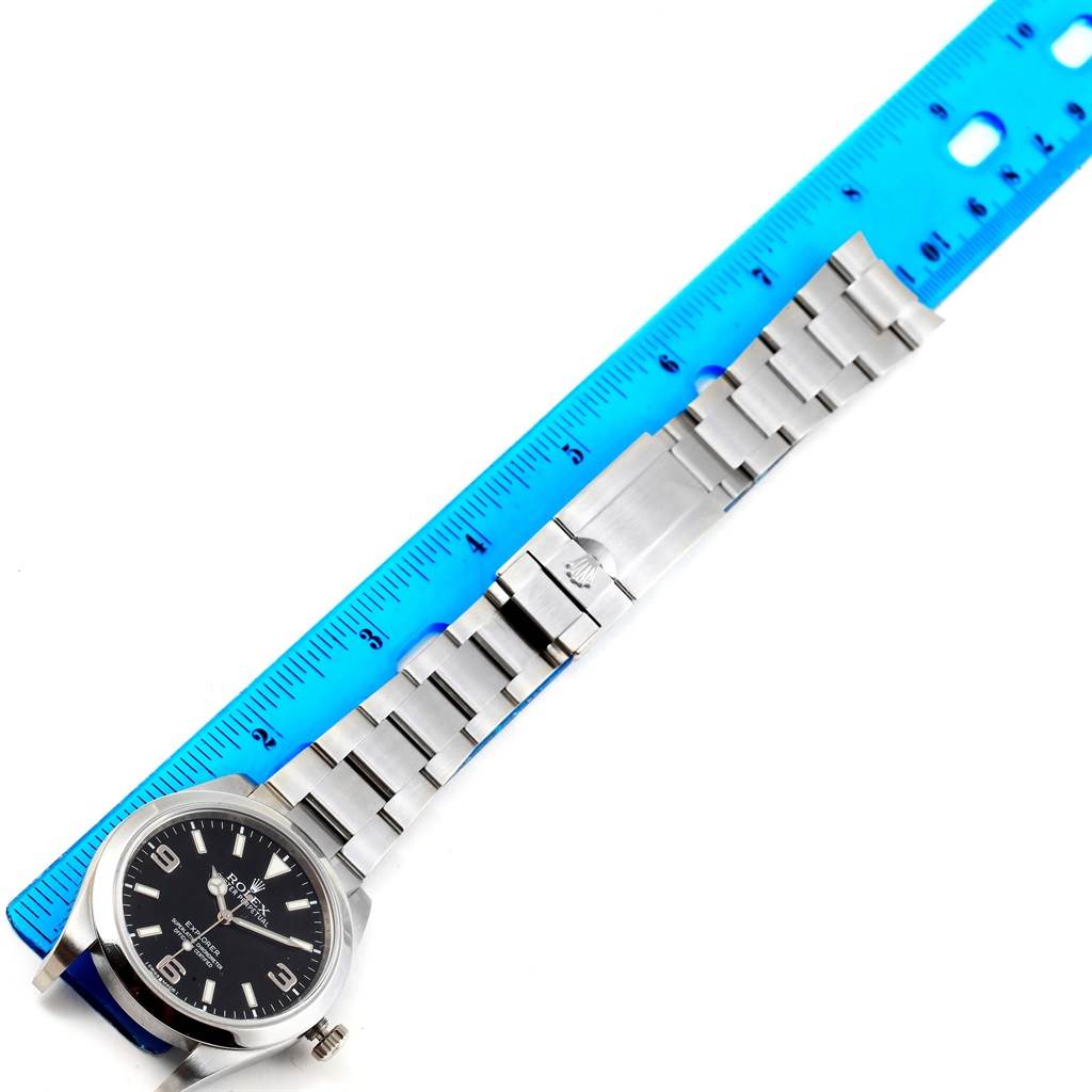 The image shows a Rolex Explorer 214270 Men's Stainless Steel Black Dial 214270 Men's Stainless Steel Black Dial watch laid flat with its bracelet extended, alongside a blue ruler for measurement.