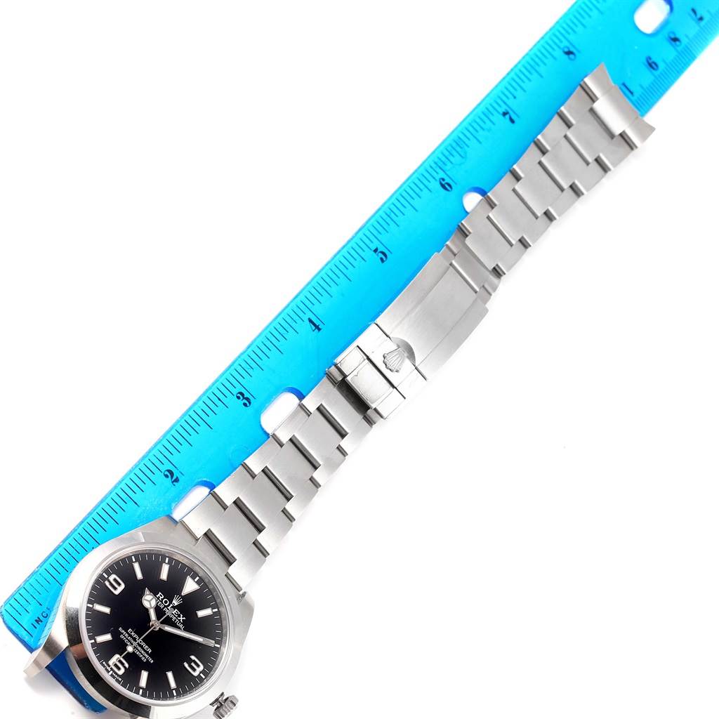 The Rolex Explorer 214270 Men's Stainless Steel Black Dial 214270 Men's Stainless Steel Black Dial watch is shown flat with its stainless steel bracelet, clasp, and part of the face visible alongside a ruler.