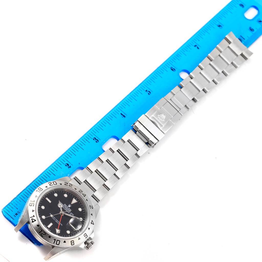 The Rolex Explorer 16677 Men's Stainless Steel Black Dial 16677 Men's Stainless Steel Black Dial watch is shown from an overhead angle, displaying its dial, bezel, and bracelet laid next to a blue ruler.