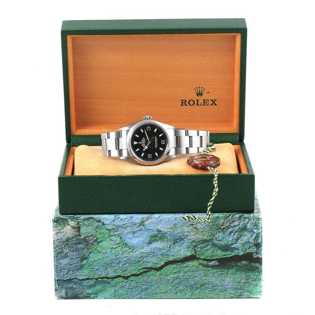 This image shows a Rolex Explorer 114270 Men's Stainless Steel Black Dial 114270 Men's Stainless Steel Black Dial watch displayed at an angle inside an open green and wooden Rolex box, with a tag accessory.