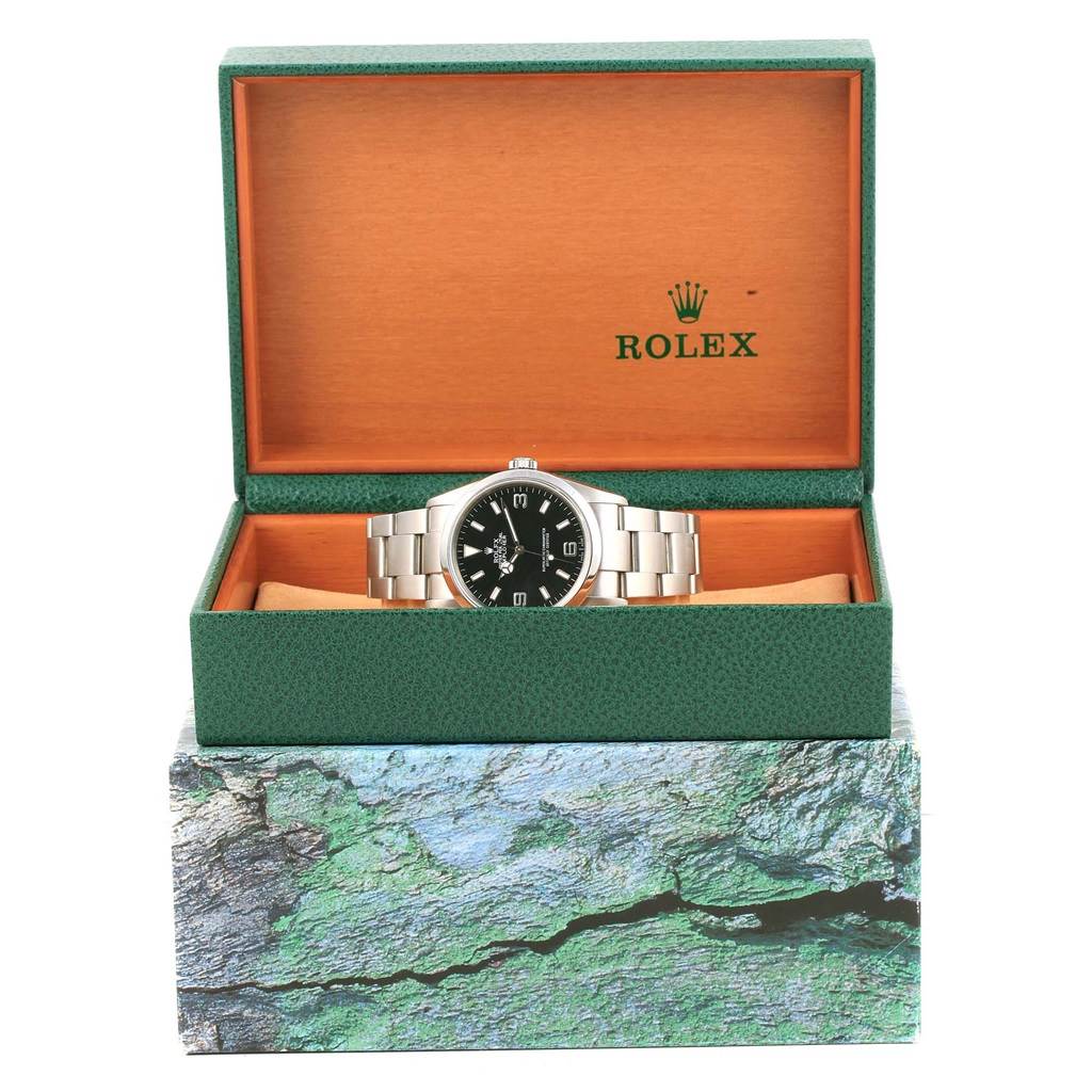 The Rolex Explorer 114270 Men's Stainless Steel Black Dial 114270 Men's Stainless Steel Black Dial watch is displayed from the front, inside its open green and orange box.
