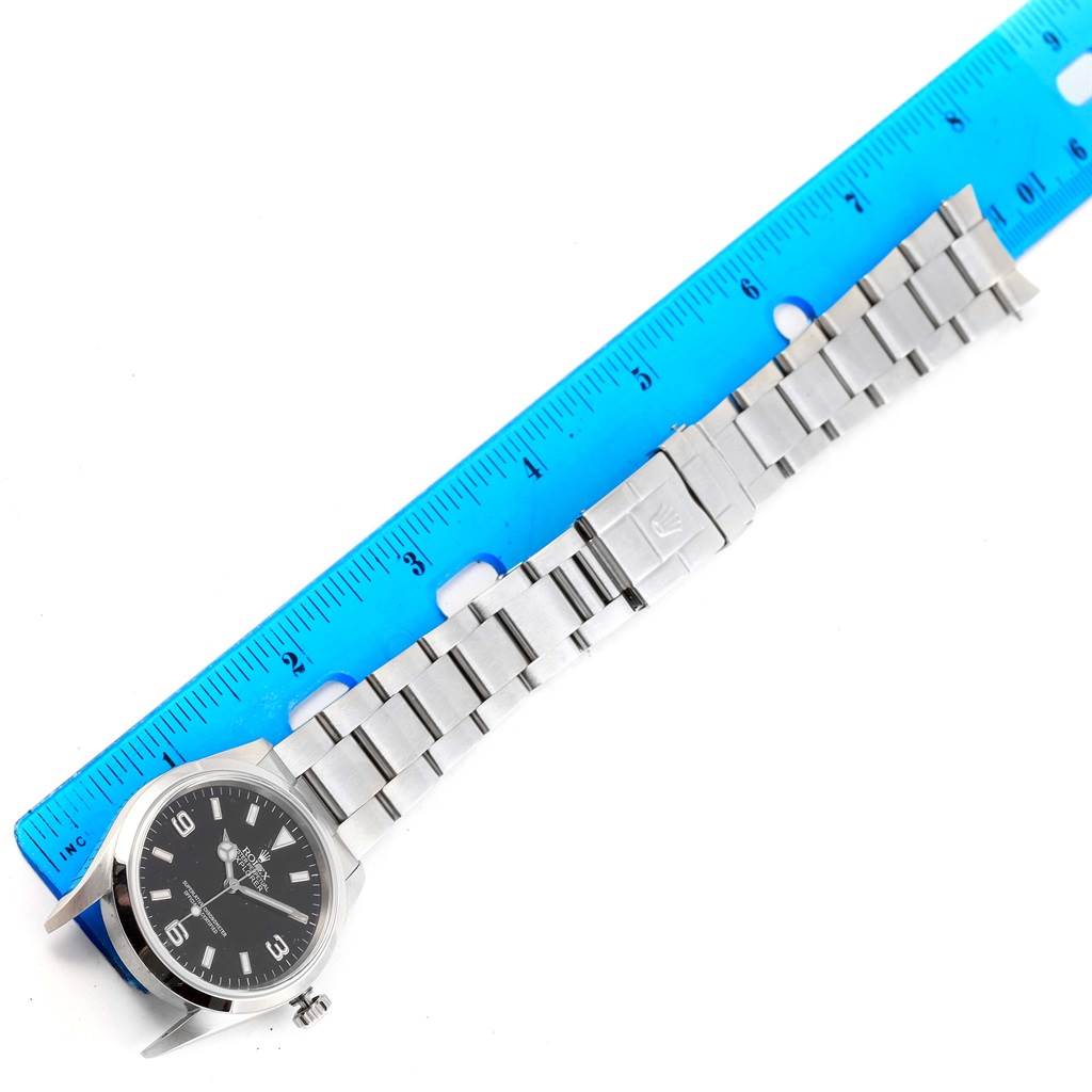 The Rolex Explorer 114270 Men's Stainless Steel Black Dial 114270 Men's Stainless Steel Black Dial watch is shown from a top-down angle, lying flat next to a blue ruler.