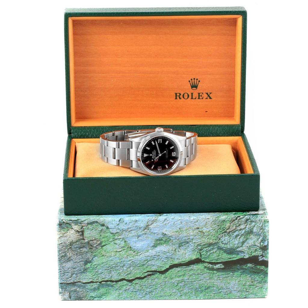 The Rolex Explorer 114270 Men's Stainless Steel Black Dial 114270 Men's Stainless Steel Black Dial watch is shown from a front angle, placed inside its open green and orange box.