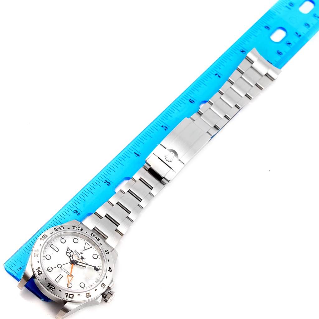 The Rolex Explorer 216570 Men's Stainless Steel White Dial 216570 Men's Stainless Steel White Dial watch is displayed at an angle showing its face, stainless steel bracelet, and clasp beside a blue ruler.