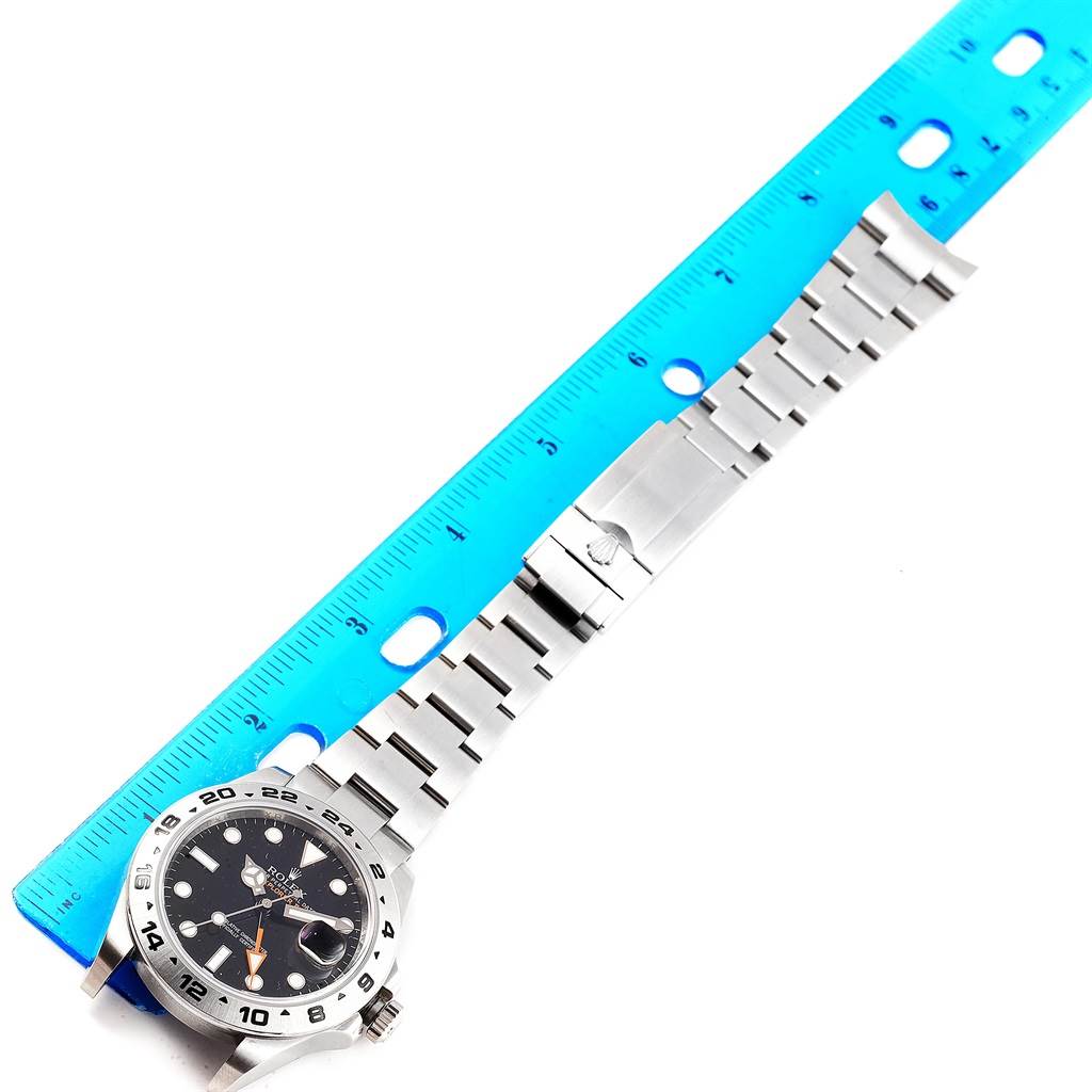 The Rolex Explorer 216570 Men's Stainless Steel Black Dial 216570 Men's Stainless Steel Black Dial watch is shown from a top-down angle with its face and stainless steel bracelet stretched alongside a blue ruler.