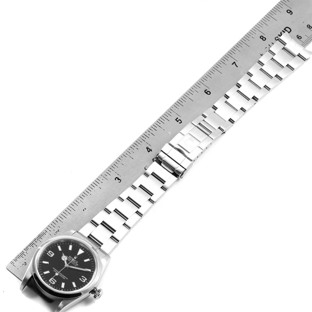 The Rolex Explorer 114270 Men's Stainless Steel Black Dial 114270 Men's Stainless Steel Black Dial watch is shown from an overhead angle along with a ruler, displaying its face, bracelet, and clasp.