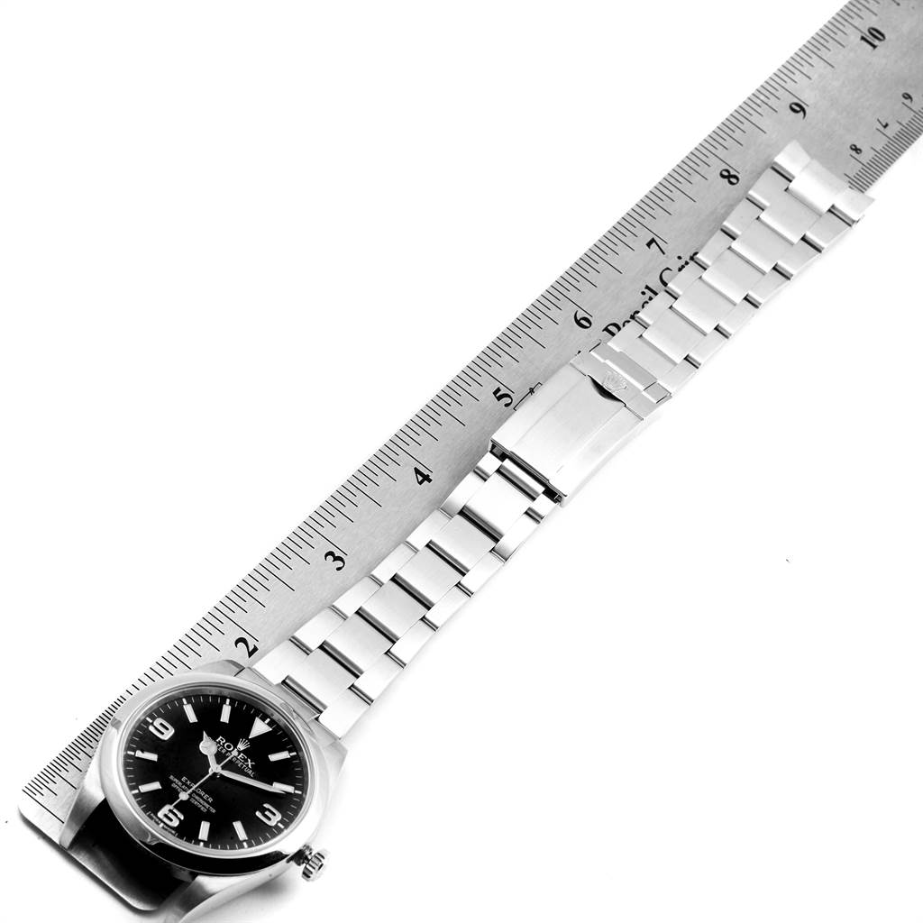 The image shows the Rolex Explorer 214270 Men
s Stainless Steel Black Dial 214270 Men
s Stainless Steel Black Dial watch laid flat alongside a ruler, displaying the watch face and the entire metal bracelet.