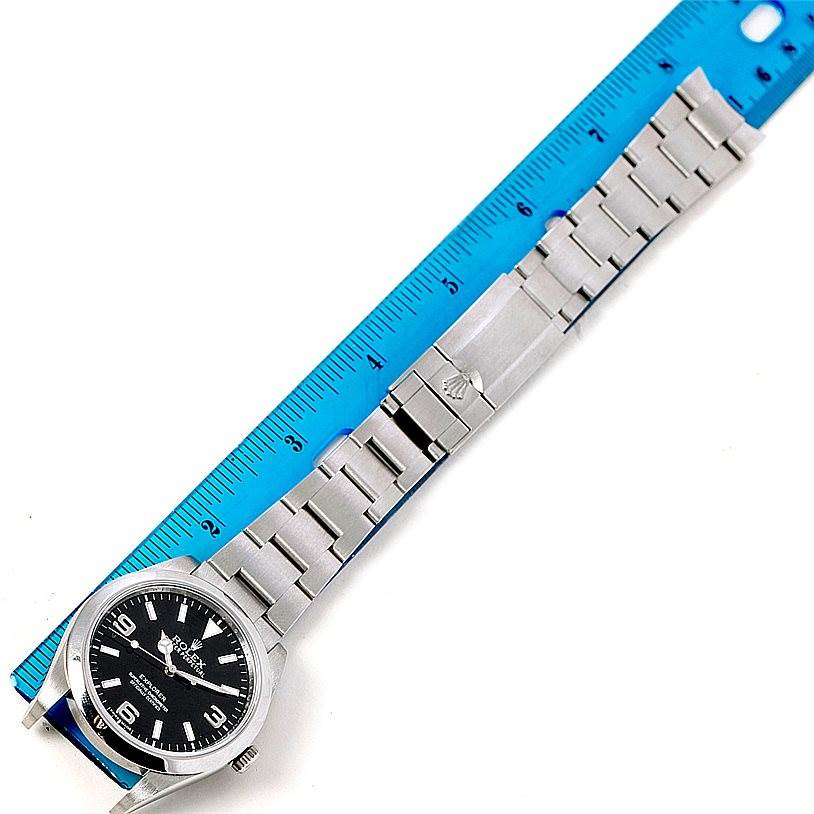 The Rolex Explorer 214270 Men's Stainless Steel Black Dial 214270 Men's Stainless Steel Black Dial watch is shown fully extended with its bracelet laid next to a blue ruler for scale.