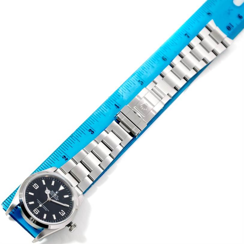 The image shows the front view of a Rolex Explorer 114270 Men's Stainless Steel Black Dial 114270 Men's Stainless Steel Black Dial watch with its metal band aligned next to a blue ruler for measurement.