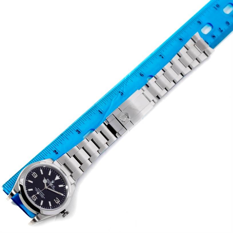 The image shows a Rolex Explorer 214270 Men's Stainless Steel Black Dial 214270 Men's Stainless Steel Black Dial watch with its dial, bracelet, and clasp, positioned next to a blue ruler for measurement.