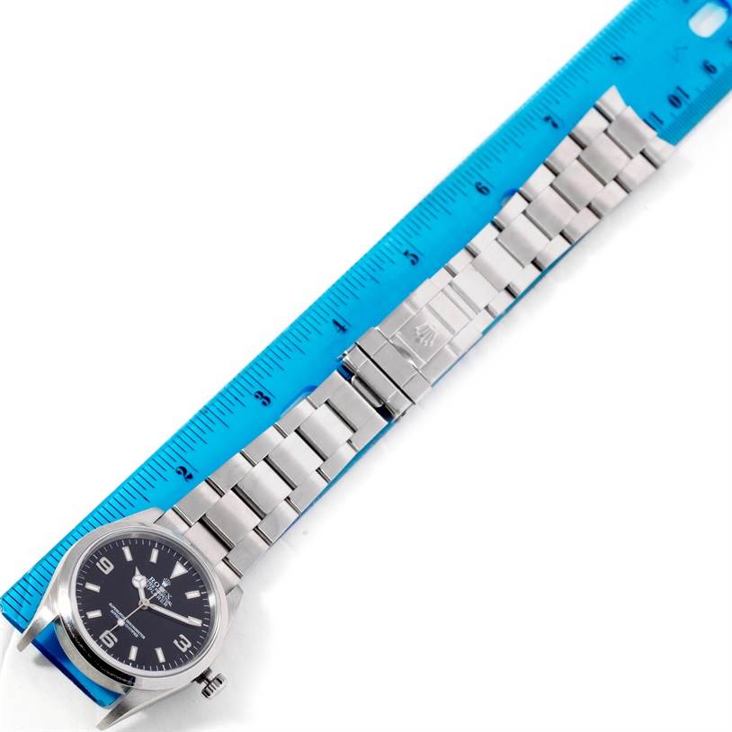 The Rolex Explorer 114270 Men's Stainless Steel Black Dial 114270 Men's Stainless Steel Black Dial watch is shown from an overhead angle, displaying its face, bracelet, and clasp against a ruler backdrop.