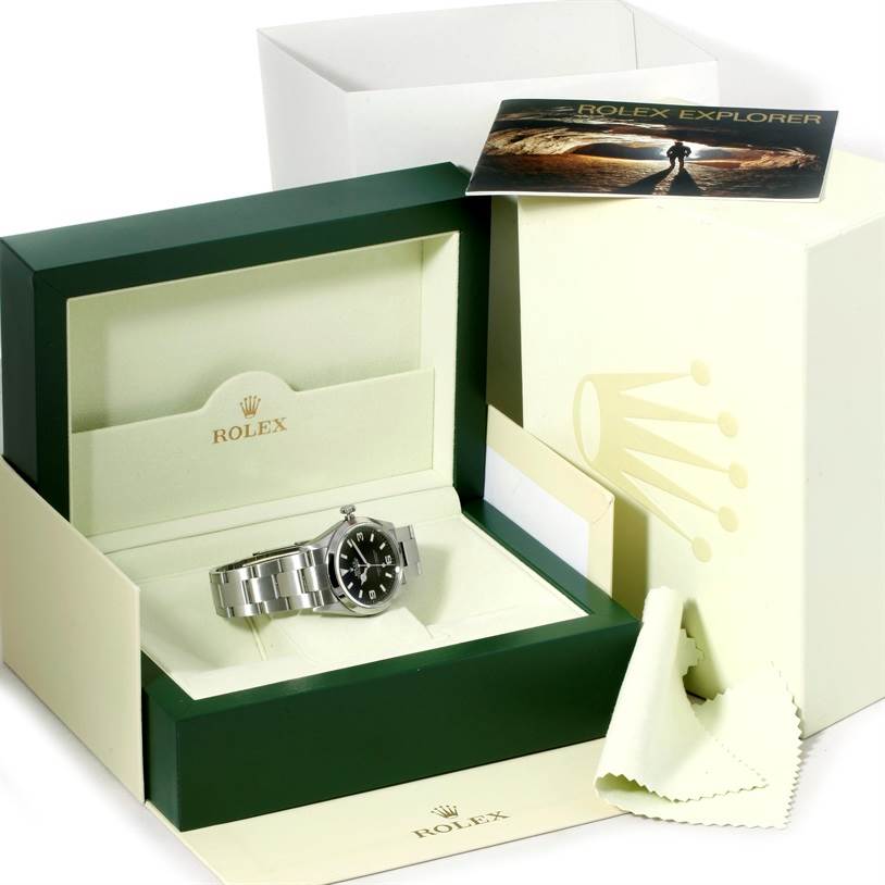 The image shows the Rolex Explorer 114270 Men's Stainless Steel Black Dial 114270 Men's Stainless Steel Black Dial watch at a frontal angle, within its box, accompanied by a booklet and a cleaning cloth.