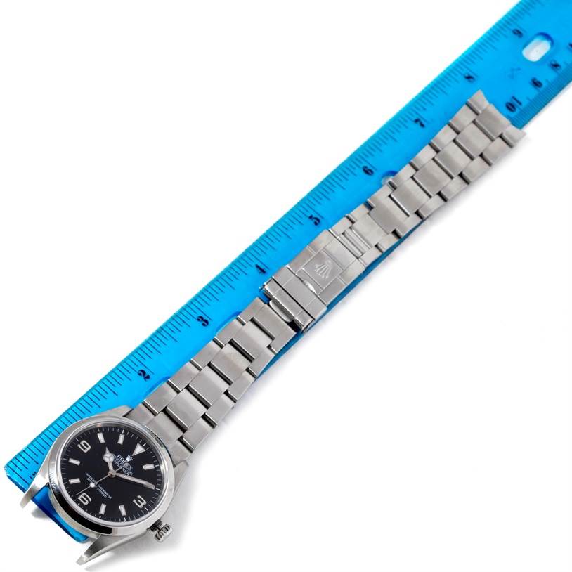 The Rolex Explorer 114270 Men's Stainless Steel Black Dial 114270 Men's Stainless Steel Black Dial watch is shown laying flat, with a prominent focus on its watch face and stainless steel bracelet alongside a blue ruler for scale.