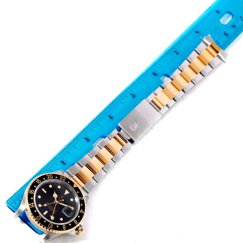 The image shows a Rolex GMT-Master 16713 Men's Stainless Steel Black Dial 16713 Men's Stainless Steel Black Dial watch laid flat next to a blue ruler, displaying its gold and silver band and black bezel.