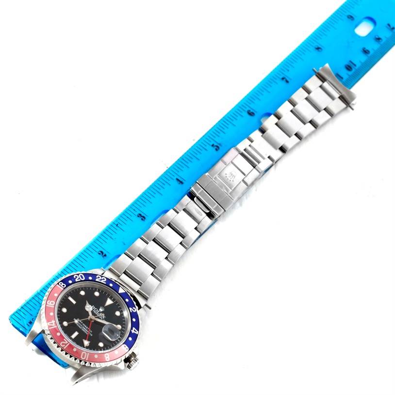 The image shows a top-down view of a Rolex GMT-Master 16700 Men's Stainless Steel Black Dial 16700 Men's Stainless Steel Black Dial watch with a red and blue bezel, positioned next to a blue ruler.