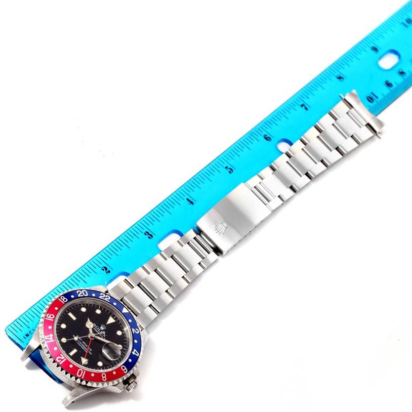 The image shows a top-down view of a Rolex GMT-Master 16700 Men's Stainless Steel Black Dial 16700 Men's Stainless Steel Black Dial watch with a blue and red bezel, next to a blue ruler for size reference.