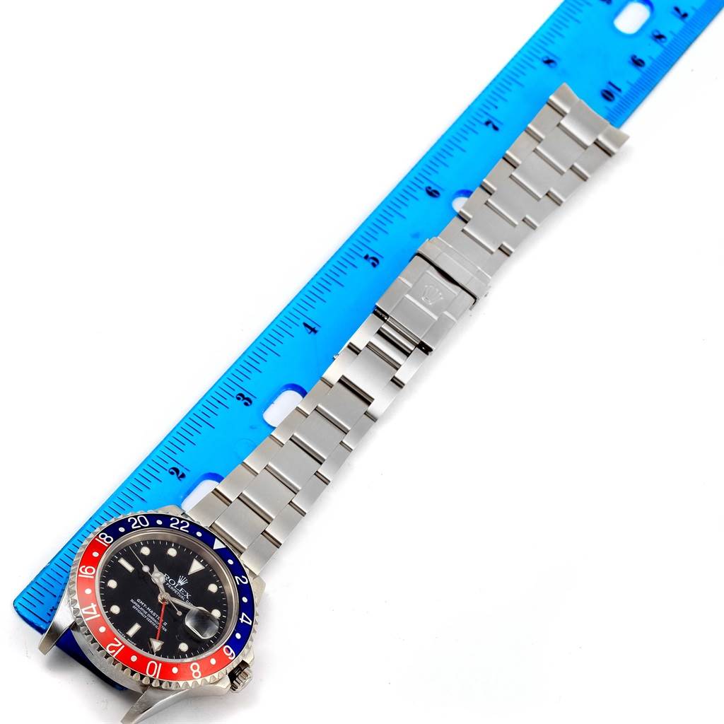The image shows the Rolex GMT-Master 16710 Men
s Stainless Steel Black Dial 16710 Men
s Stainless Steel Black Dial watch and its entire stainless steel bracelet positioned alongside a blue ruler.