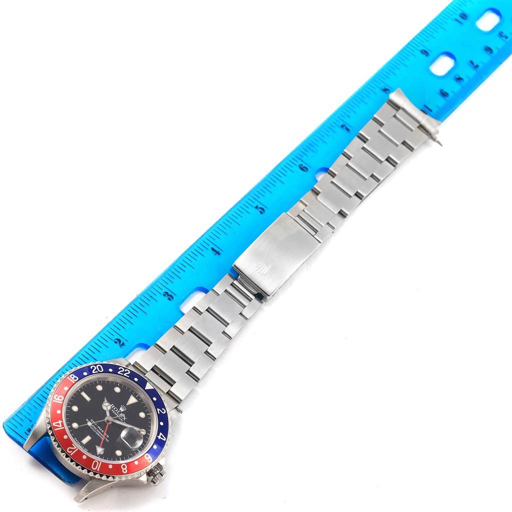 The image shows a full view of a Rolex GMT-Master 16700 Men
s Stainless Steel Black Dial 16700 Men
s Stainless Steel Black Dial watch lying flat with its bracelet extended next to a blue ruler.