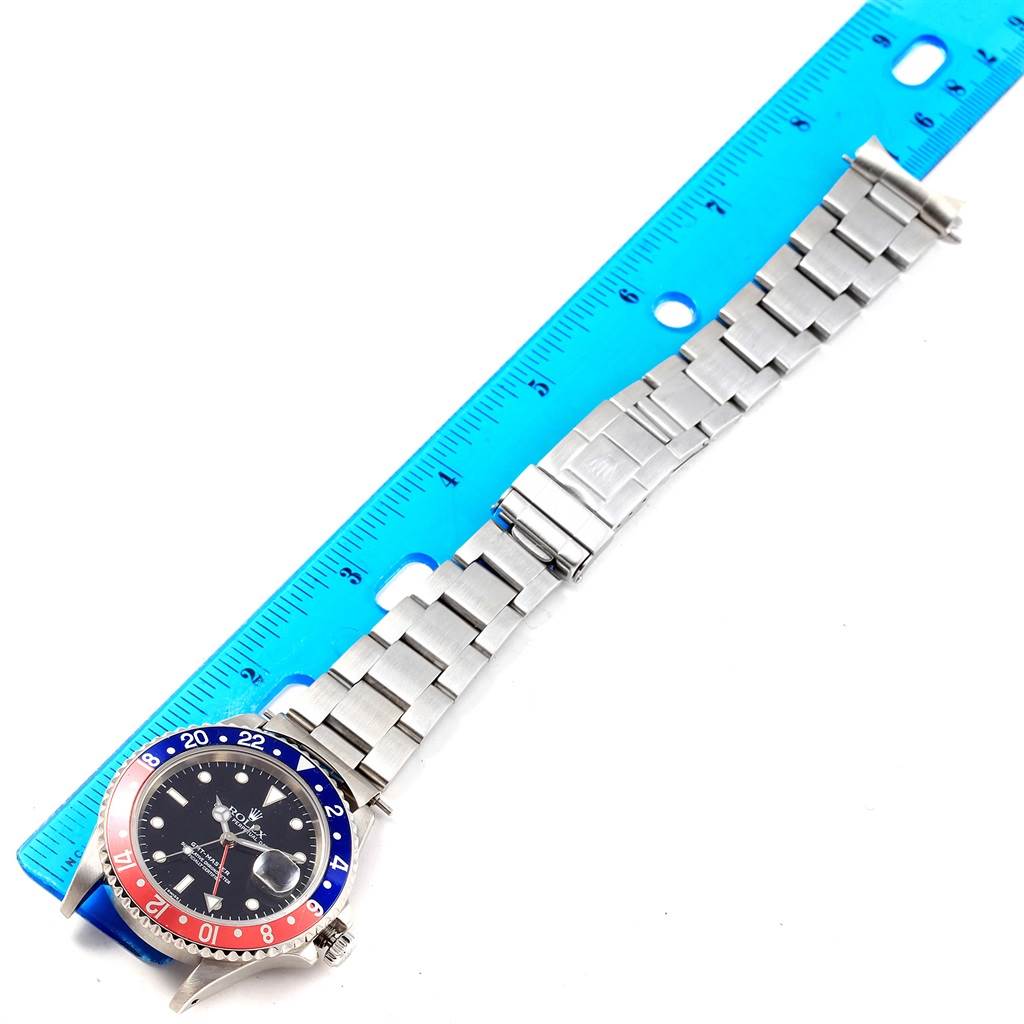 The image shows a flat, top-down view of a Rolex GMT-Master 16700 Men's Stainless Steel Black Dial 16700 Men's Stainless Steel Black Dial watch with its stainless steel bracelet and a blue and red bezel, next to a blue ruler.