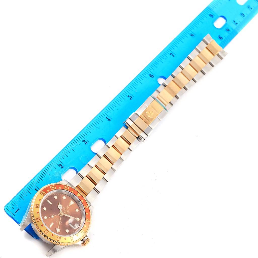 The Rolex GMT-Master 16713 Men's Stainless Steel Brown Dial 16713 Men's Stainless Steel Brown Dial watch is shown from the top view along with its band, positioned beside a blue ruler.