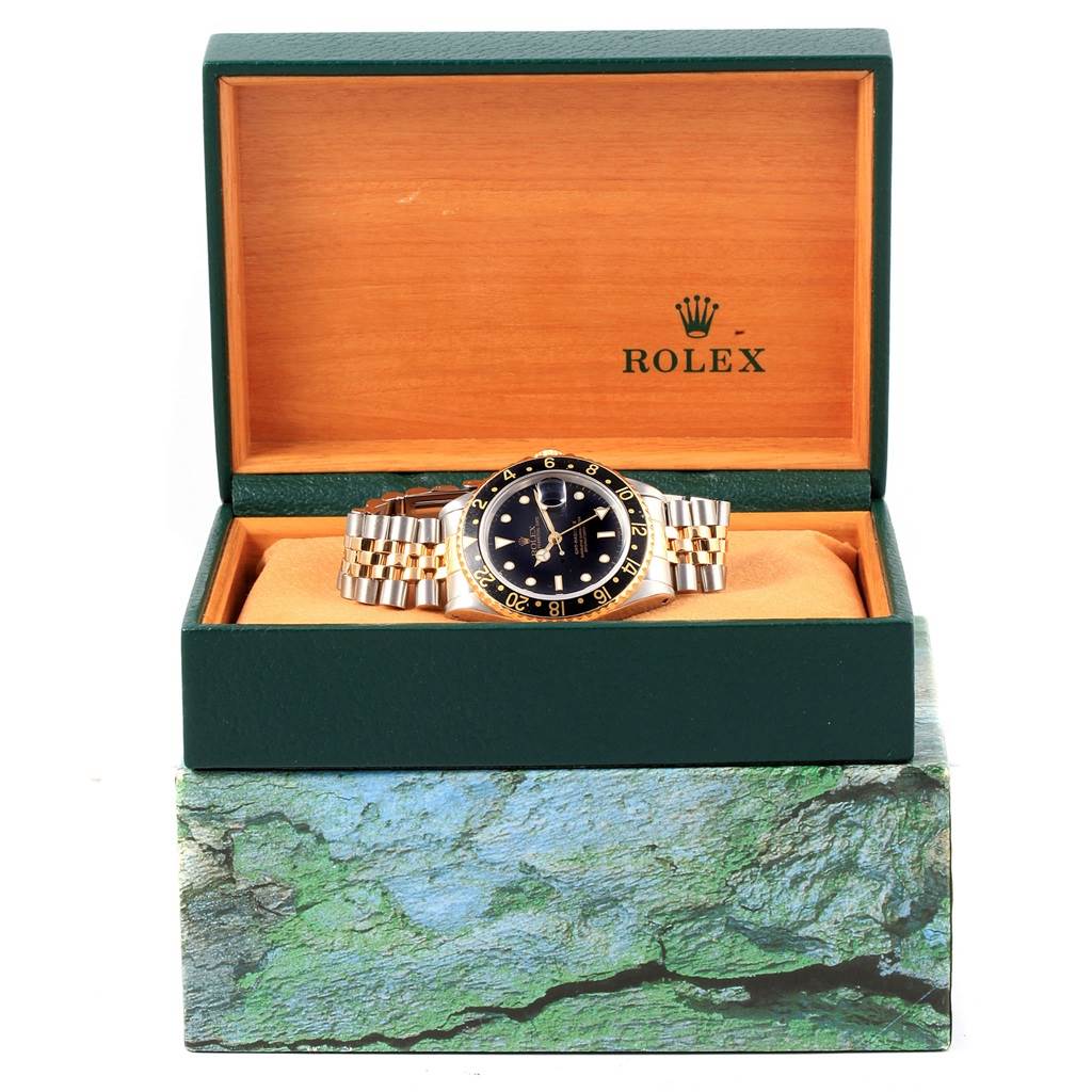 The Rolex GMT-Master 16713 Men's Stainless Steel Black Dial 16713 Men's Stainless Steel Black Dial watch is displayed front-on in a green and wood-patterned box.
