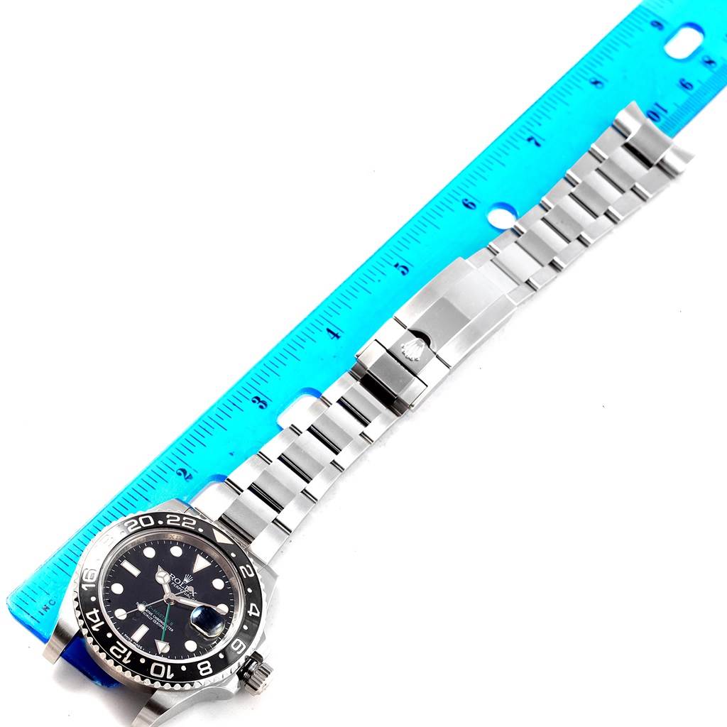 The image shows a GMT-Master Rolex watch laid flat with a blue ruler beside it, highlighting the bezel, dial, and stainless steel bracelet.