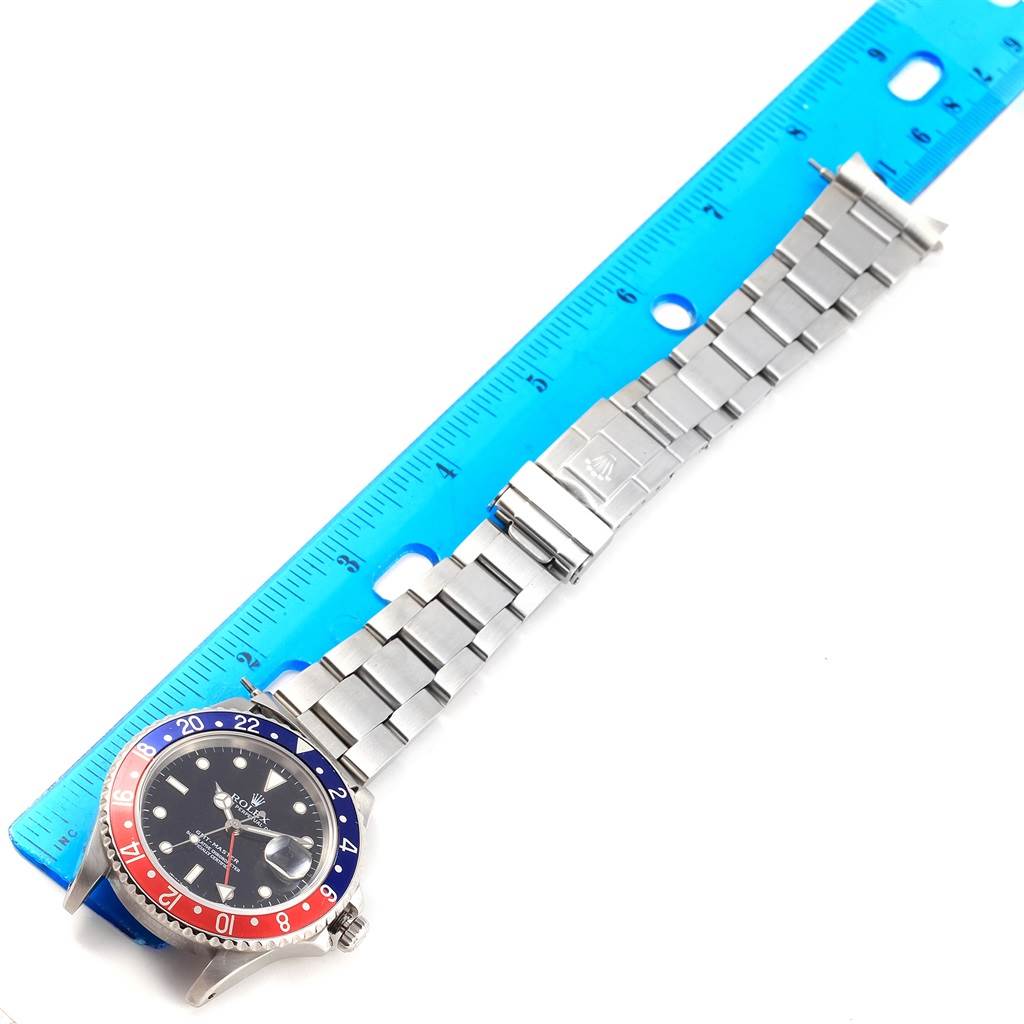 The image shows a top-down view of the Rolex GMT-Master 16700 Men's Stainless Steel Black Dial 16700 Men's Stainless Steel Black Dial watch, featuring its bracelet and the "Pepsi" bezel, next to a blue ruler.