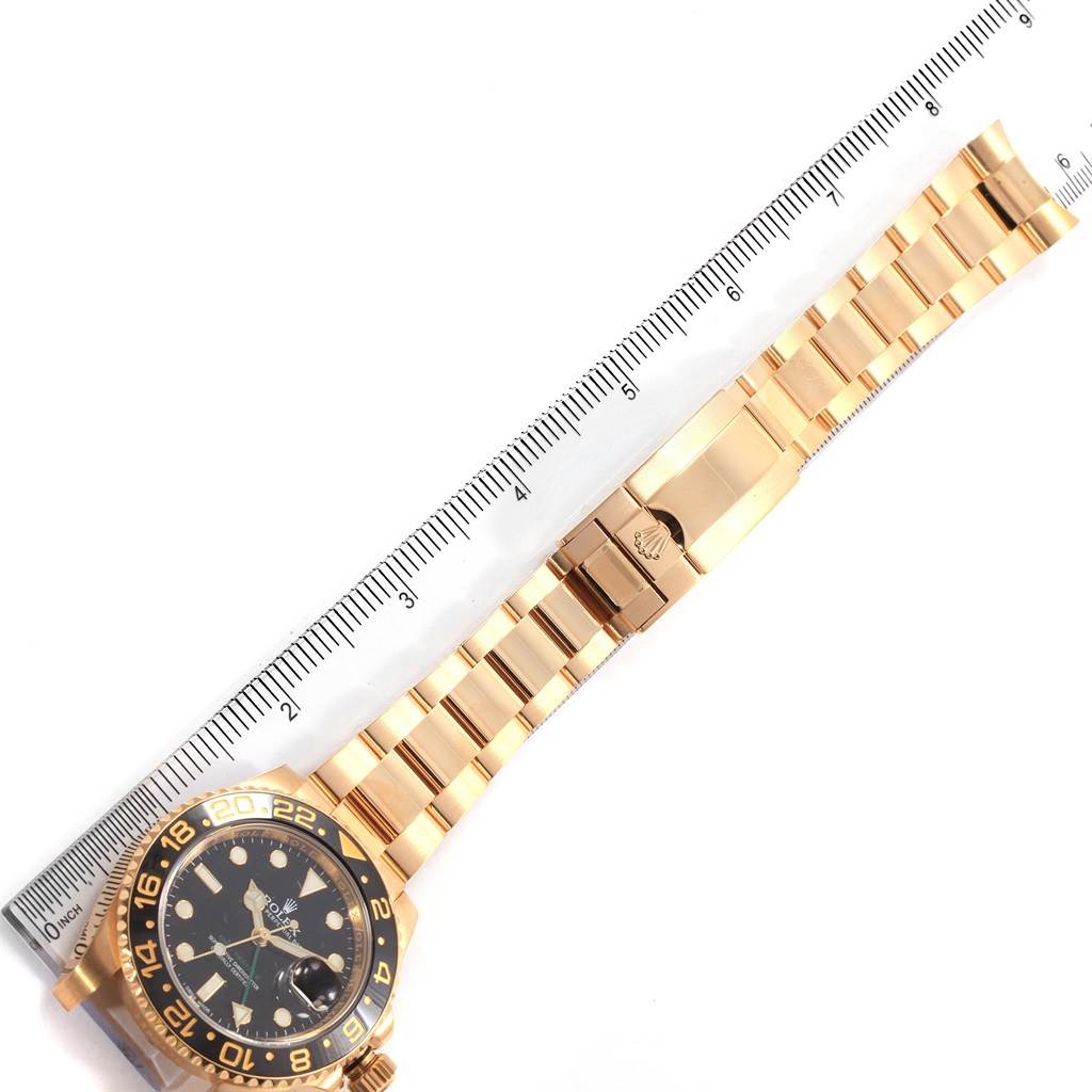 This image shows a side view of a Rolex GMT-Master 116718 Men
s Yellow Gold Black Dial 116718 Men
s Yellow Gold Black Dial watch with a gold bracelet laid out beside a ruler.