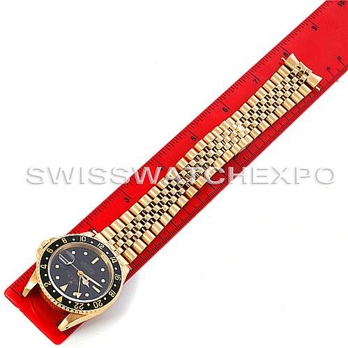 The image shows a top view of a Rolex GMT-Master 16758 Men's Stainless Steel Black Dial 16758 Men's Stainless Steel Black Dial watch with a gold bracelet alongside a red measuring ruler.