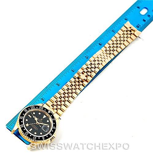 This image shows the face and full bracelet of a Rolex GMT-Master 16718 Men's Yellow Gold Black Dial 16718 Men's Yellow Gold Black Dial watch placed next to a blue measuring ruler.
