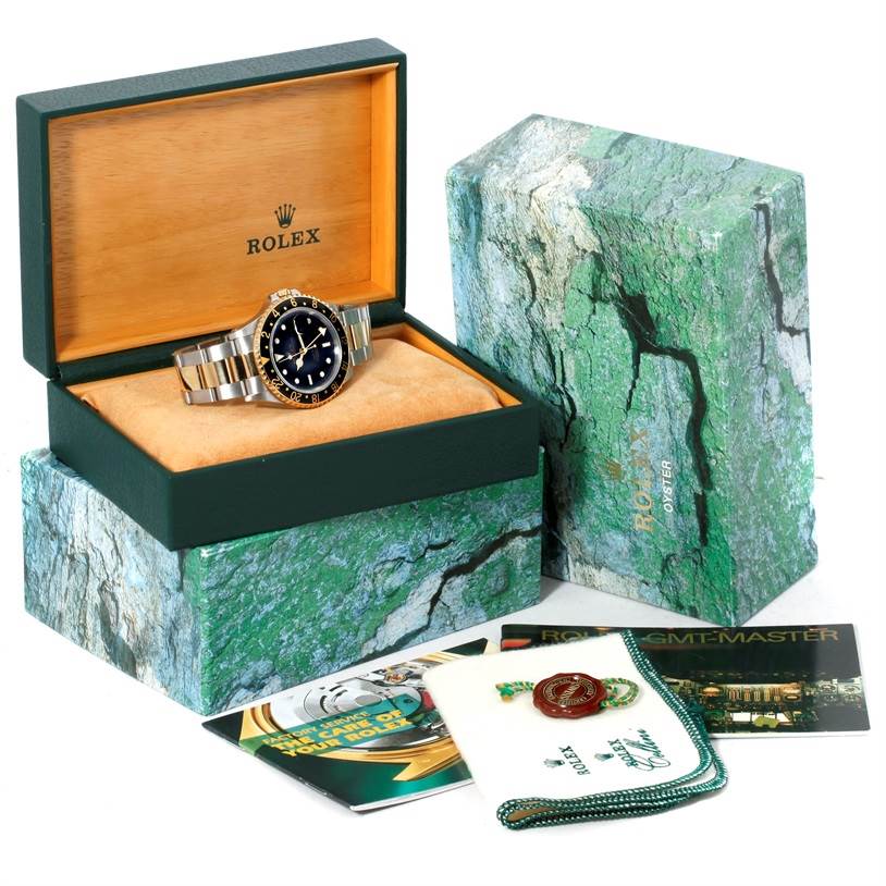 The image shows a Rolex GMT-Master 16713 Men
s Stainless Steel Black Dial 16713 Men
s Stainless Steel Black Dial watch in an open green box with additional accessories and documentation around it.