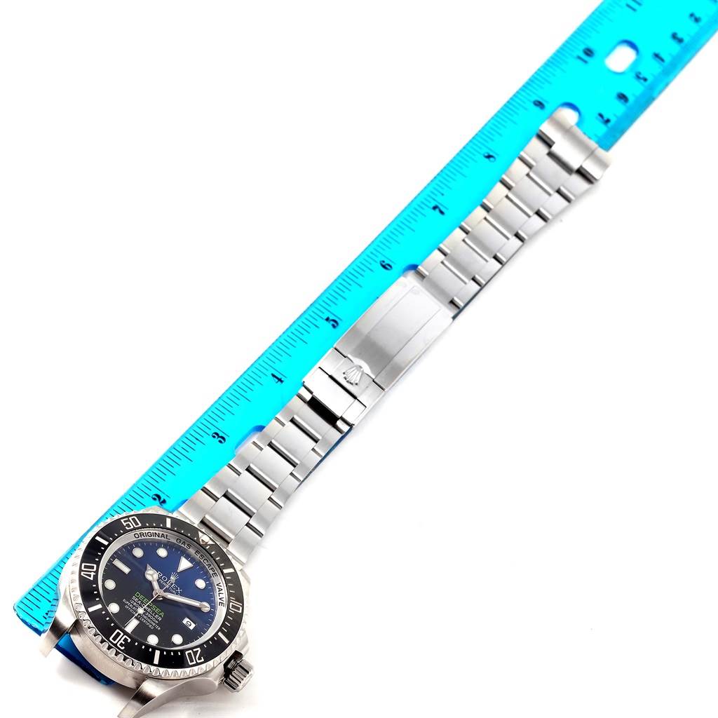 The Rolex Sea-Dweller 116660 Men
s Stainless Steel Blue Dial 116660 Men
s Stainless Steel Blue Dial watch is shown laid flat, displaying the face, bezel, bracelet, and clasp next to a blue ruler.