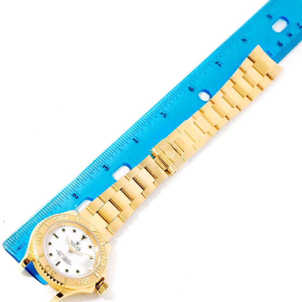 The image shows the front view of a Rolex Yacht-Master 16628 Men's Yellow Gold White Dial 16628 Men's Yellow Gold White Dial watch with a gold case and bracelet beside a blue ruler.