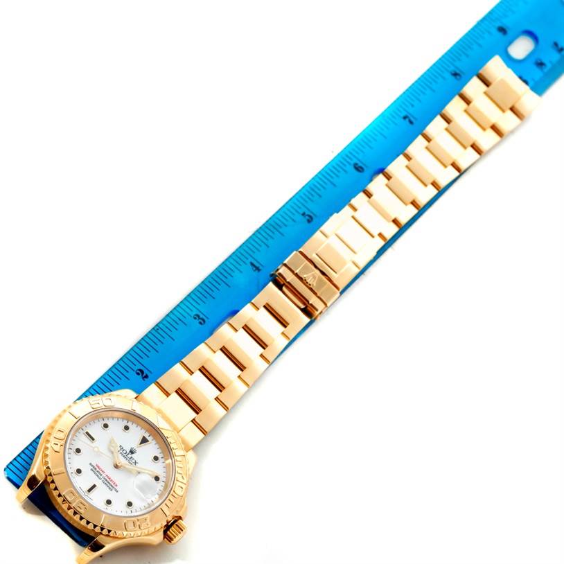 The image shows a top view of a Rolex Yacht-Master 16628 Men's Yellow Gold White Dial 16628 Men's Yellow Gold White Dial watch with its entire bracelet and face aligned next to a ruler.