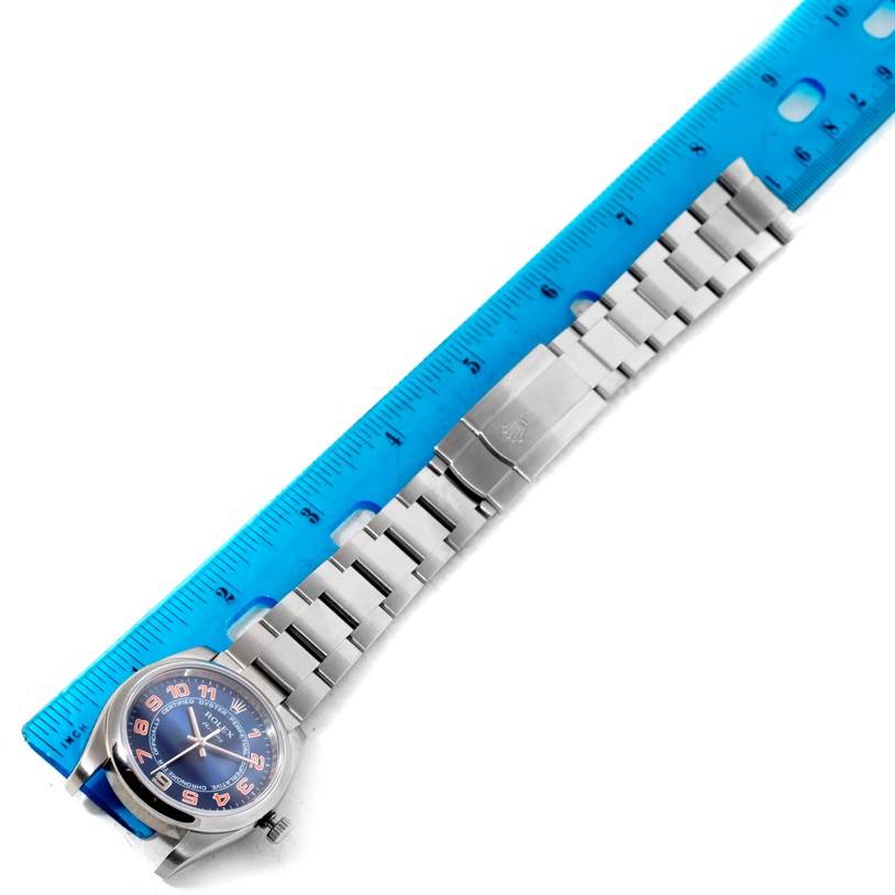 The image shows a top-down view of a Rolex Air-King 114200 Men
s Stainless Steel Blue Dial 114200 Men
s Stainless Steel Blue Dial watch with its bracelet aligned beside a blue ruler.