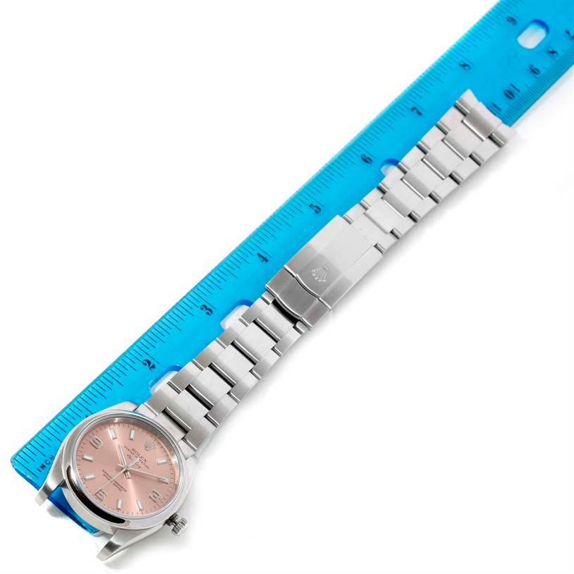 The Rolex Air-King 114200 Men's Stainless Steel Pink Dial 114200 Men's Stainless Steel Pink Dial watch is shown from a top-down angle, displaying its bracelet, clasp, and face next to a blue ruler.