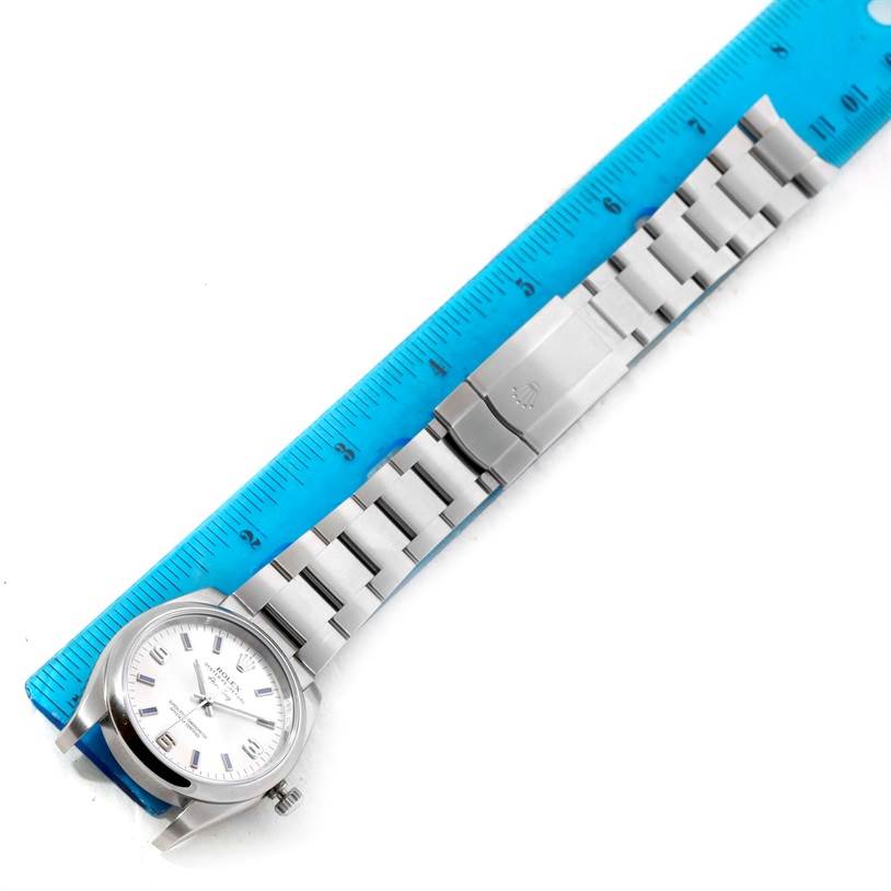 The Rolex Air-King 114200 Men's Stainless Steel Silver Dial 114200 Men's Stainless Steel Silver Dial watch is shown flat, with its face, bracelet, and clasp visible alongside a blue ruler.