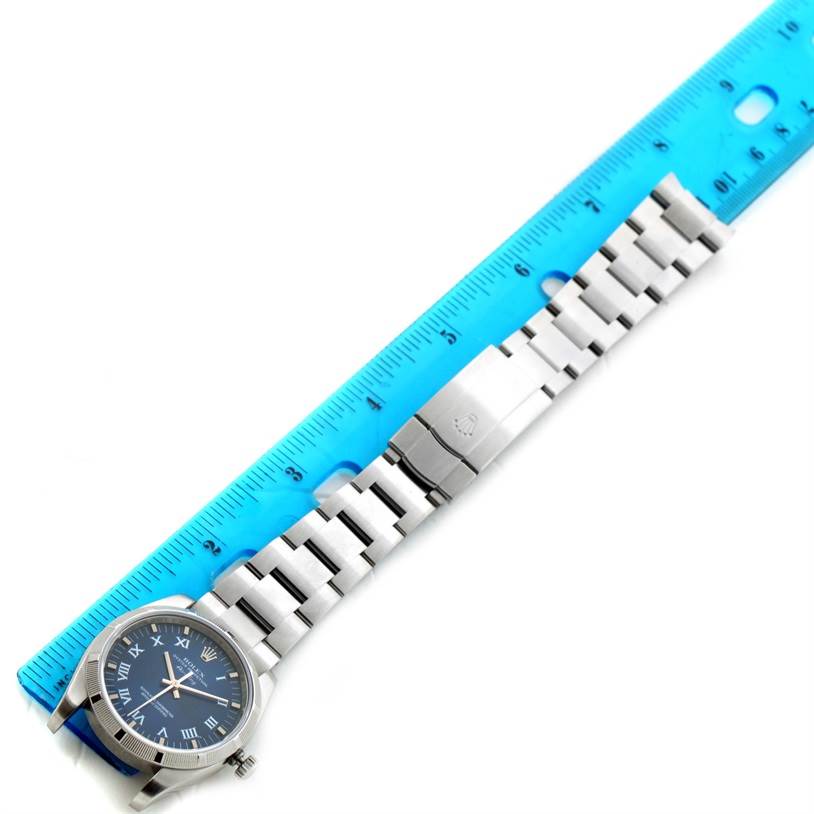 The Rolex Air-King 114200BLRO Men's Stainless Steel Blue Dial 114200BLRO Men's Stainless Steel Blue Dial watch is shown from a top-down angle, displaying the face, bracelet, and clasp next to a blue ruler.