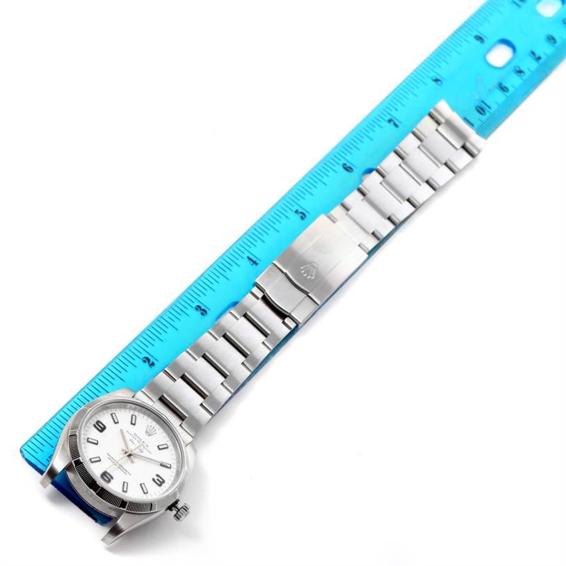 The Rolex Air-King 114210 Men's Stainless Steel White Dial 114210 Men's Stainless Steel White Dial watch is shown flat on its face with the full band extended alongside a blue ruler.