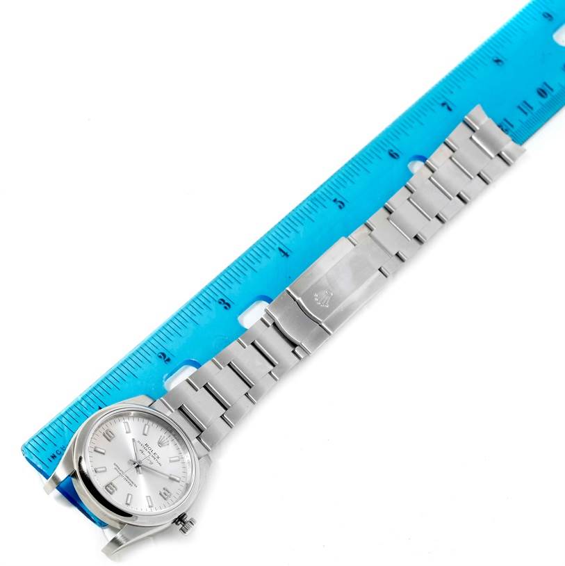 The image shows a top-down view of a Rolex Air-King 114200 Men's Stainless Steel Silver Dial 114200 Men's Stainless Steel Silver Dial watch next to a blue ruler, highlighting its dial, bracelet, and clasp.