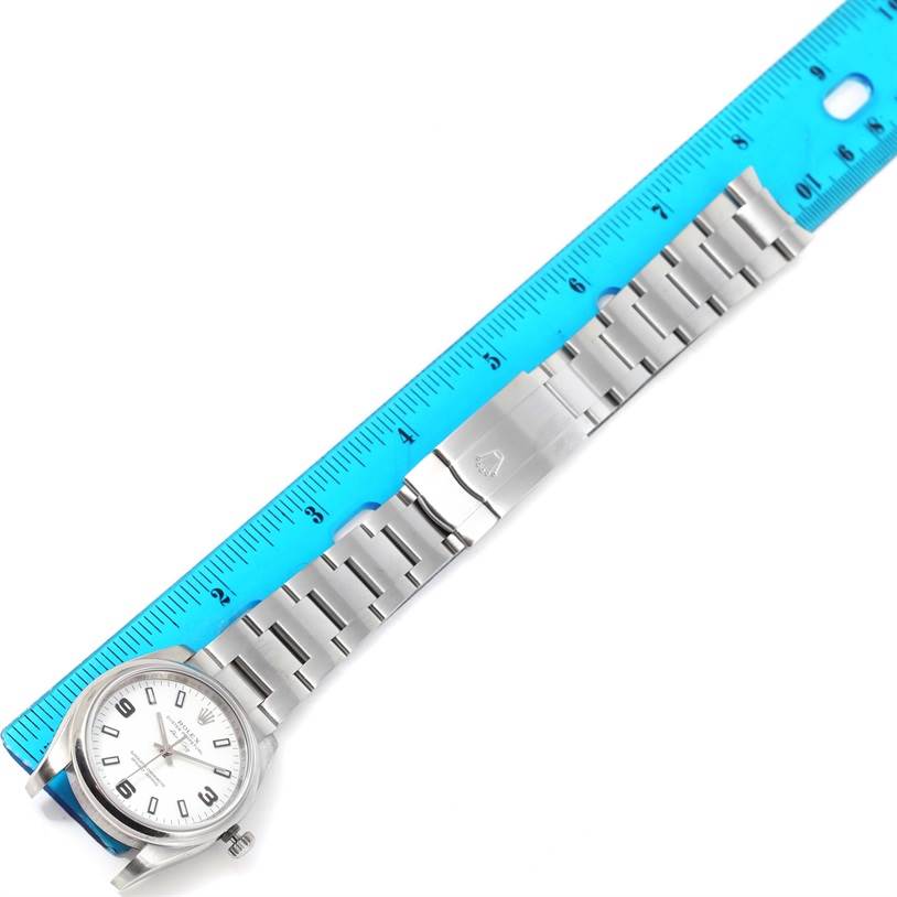 The Rolex Air-King 114200 Men's Stainless Steel White Dial 114200 Men's Stainless Steel White Dial watch is shown from a top-down angle, with its stainless steel bracelet laid out beside a blue ruler.