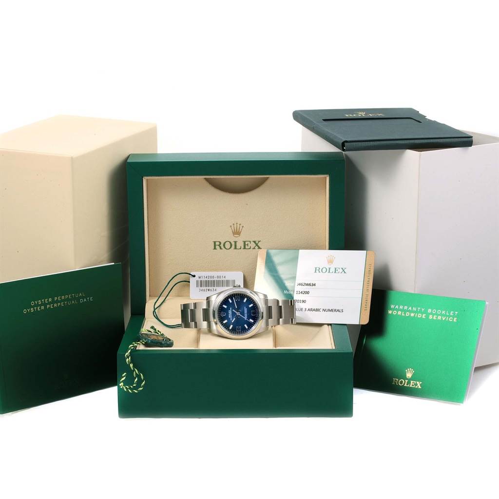 The Rolex Air-King 114200 Men's Stainless Steel Blue Dial 114200 Men's Stainless Steel Blue Dial watch is displayed front-on in its box, with warranty cards and booklets surrounding it.